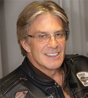 A middle-aged man in a lather jacket and glasses smiling
